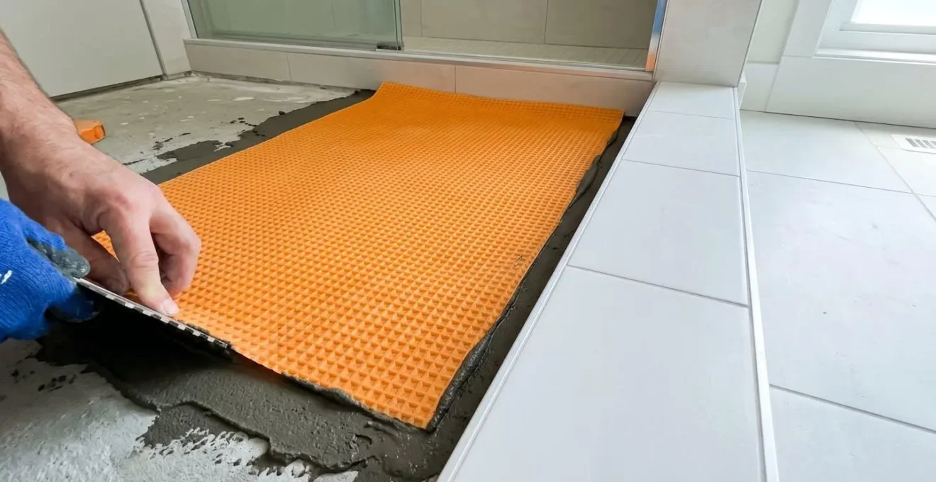 Gros plan sur une membrane d'étanchéité de couleur orange en cours d'application sur le plancher d'une douche avec truelle visible