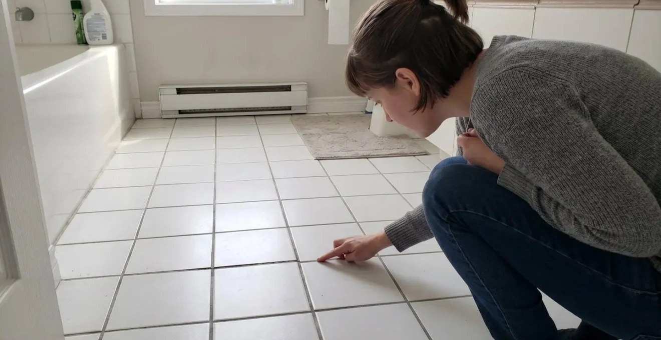Personne de profil accroupie inspectant avec le doigt un joint de céramique au plancher d'une salle de bain résidentielle contemporaine